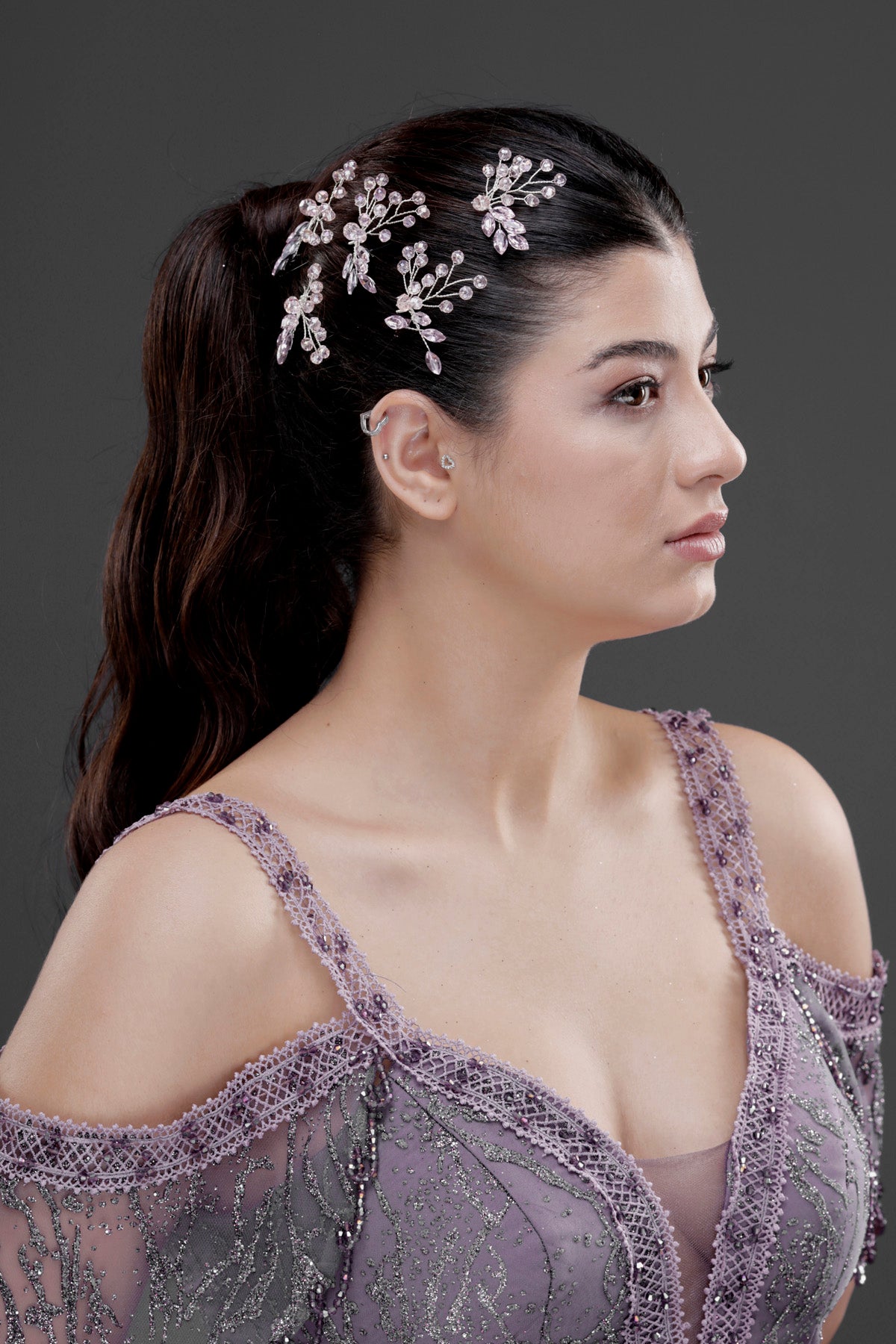 Gümüş kristal çiçekli dal saç aksesuarı – yarı topuz saç modelinde yan profil görünümü, lila dantel soğuk omuz elbise ile uyumlu sofistike düğün ve nişan saç süsü
