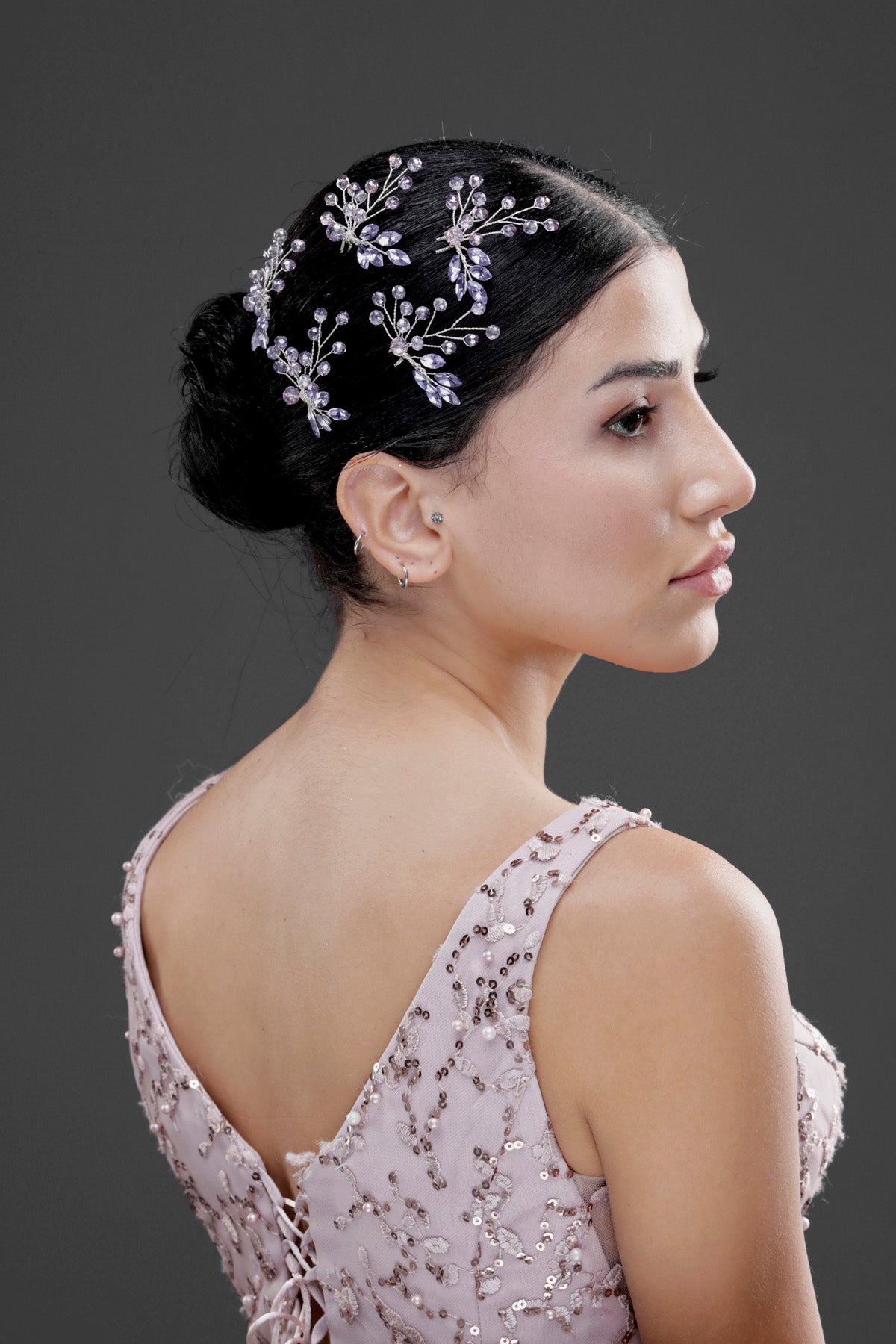 Gümüş kristal dallı çok küme saç aksesuarı – koyu topuzlu saç modelinde yan görünüm, pudra pembe çiçek nakışlı sırt dekolteli gelinlik ile uyumlu sofistike nikah saç süsü
